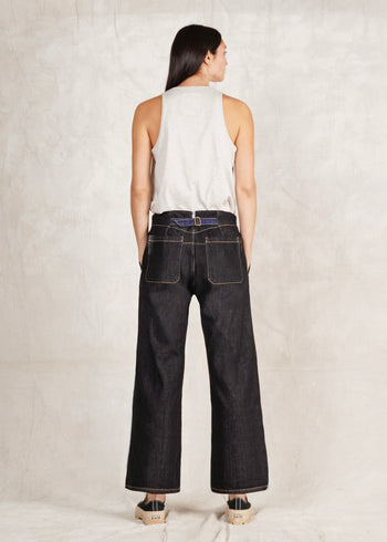 A female model wearing a white sleeveless top and blue denim artist pants with a high waist cut. She is standing against a plain background with back to us