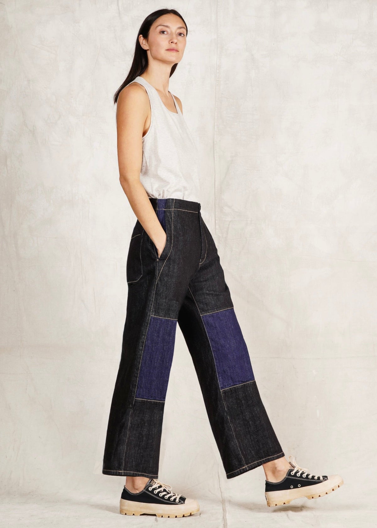 A female model wearing a white sleeveless top and blue denim artist pants with a high waist cut. She is sideways against a plain background.