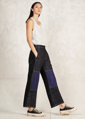 A female model wearing a white sleeveless top and blue denim artist pants with a high waist cut. She is sideways against a plain background.