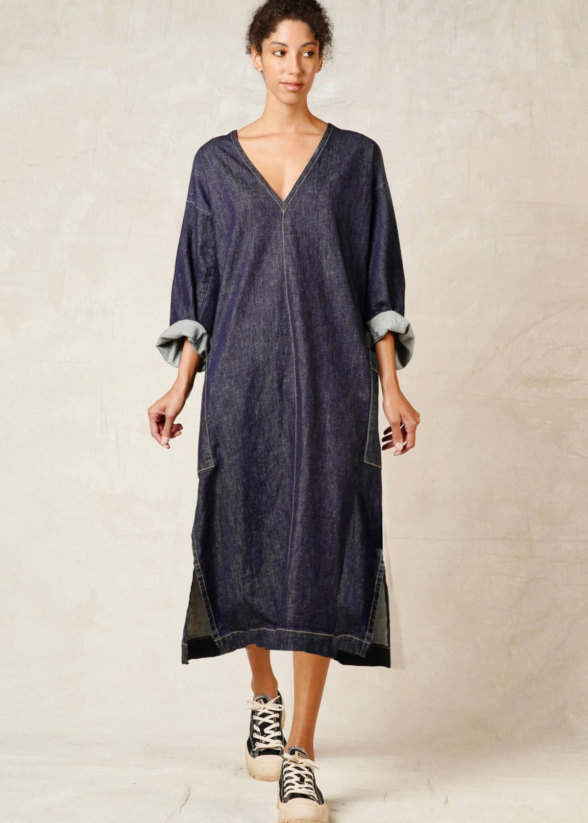 woman in v-neck loose fit denim dress with rolled sleeves and sneakers against neutral wall