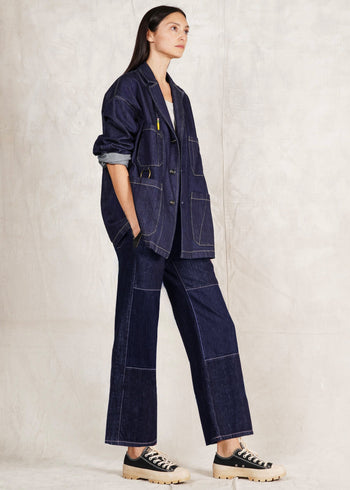 Woman wearing a dark denim Worker's jacket and Artist pants against a plain background