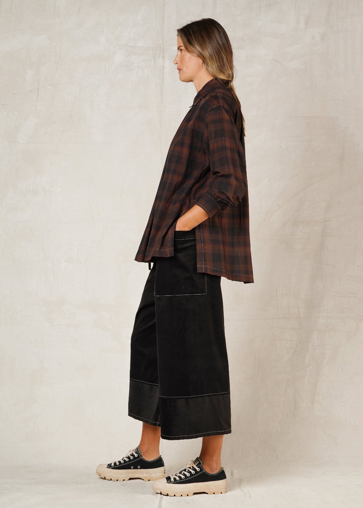 side view of model on a plain background, wearing a long-sleeved checkered shirt with a collar and high-low hem, paired with black trousers, hand in pocket and casual shoes.