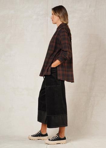 side view of model on a plain background, wearing a long-sleeved checkered shirt with a collar and high-low hem, paired with black trousers, hand in pocket and casual shoes.