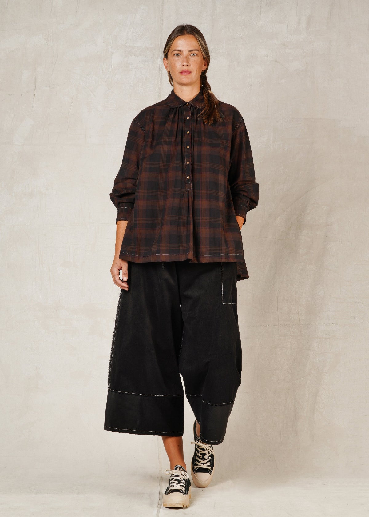 A woman standing on a plain background, wearing a long-sleeved checkered shirt with a collar and high-low hem, paired with black trousers and casual shoes.