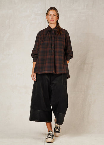 A person standing on a plain background, wearing a long-sleeved checkered shirt with a collar and high-low hem, paired with black trousers and casual shoes.