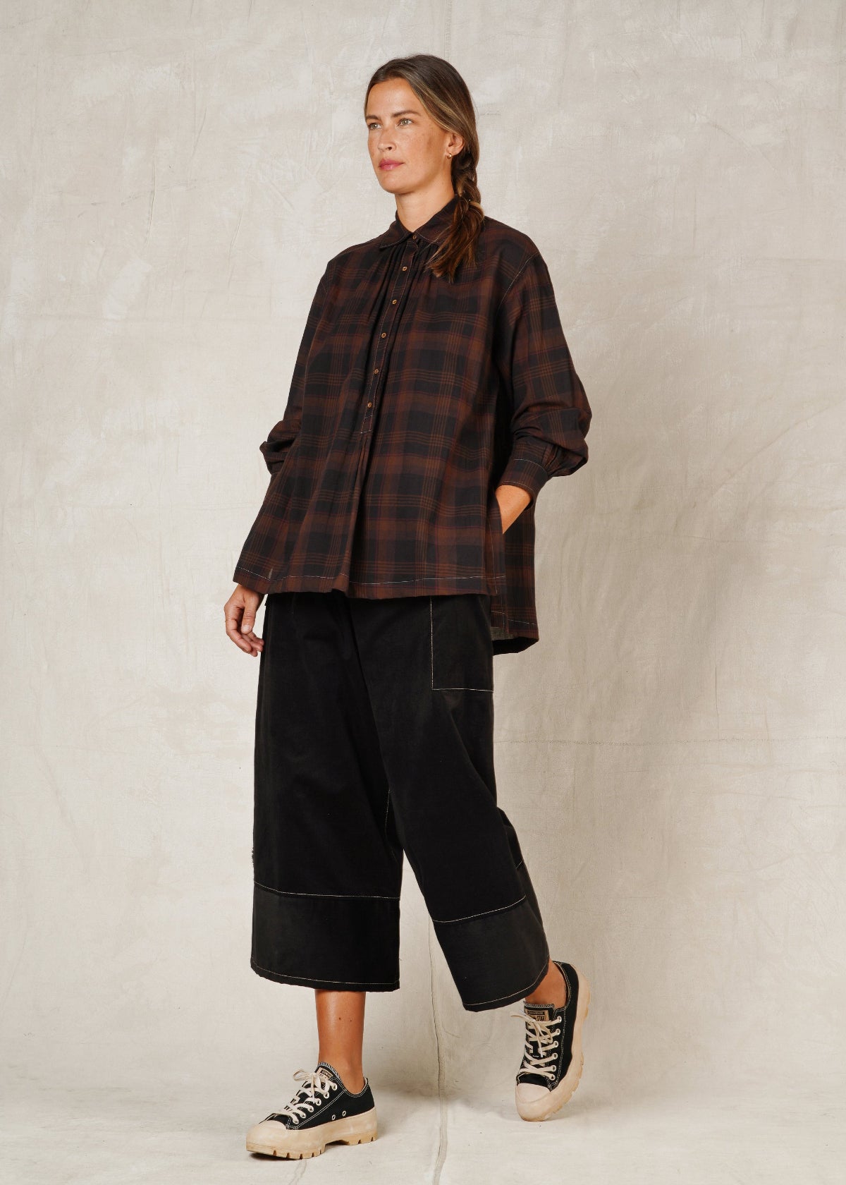 A woman standing on a plain background, wearing a long-sleeved checkered shirt with a collar and high-low hem, paired with black trousers and casual shoes.